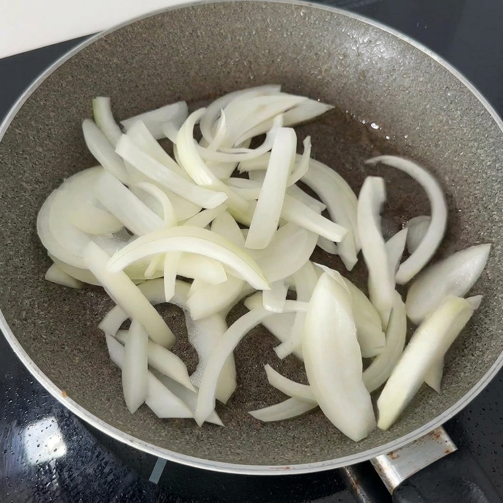 Thinly sliced white onions being stir-fried in a dark pan with residual oil until they become soft.