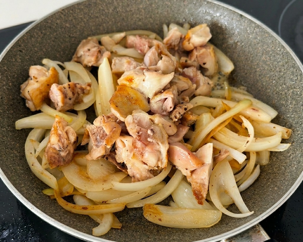 Sautéed onion slices mixed with browned chicken thigh pieces and green onions in a non-stick frying pan.