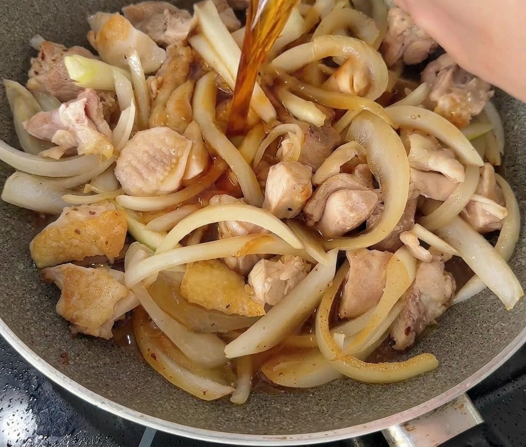 A dark, savory liquid being poured from a white bowl into a pan filled with chicken and onion slices.