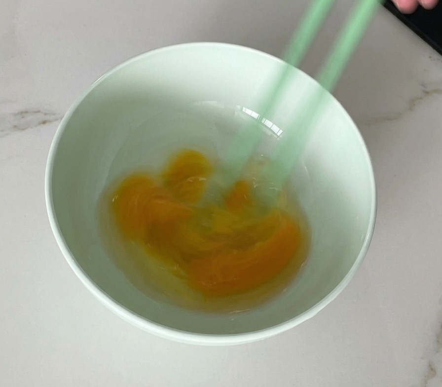 A pair of green chopsticks whisking raw eggs in a light green bowl, showing a marbled yellow and white mixture.