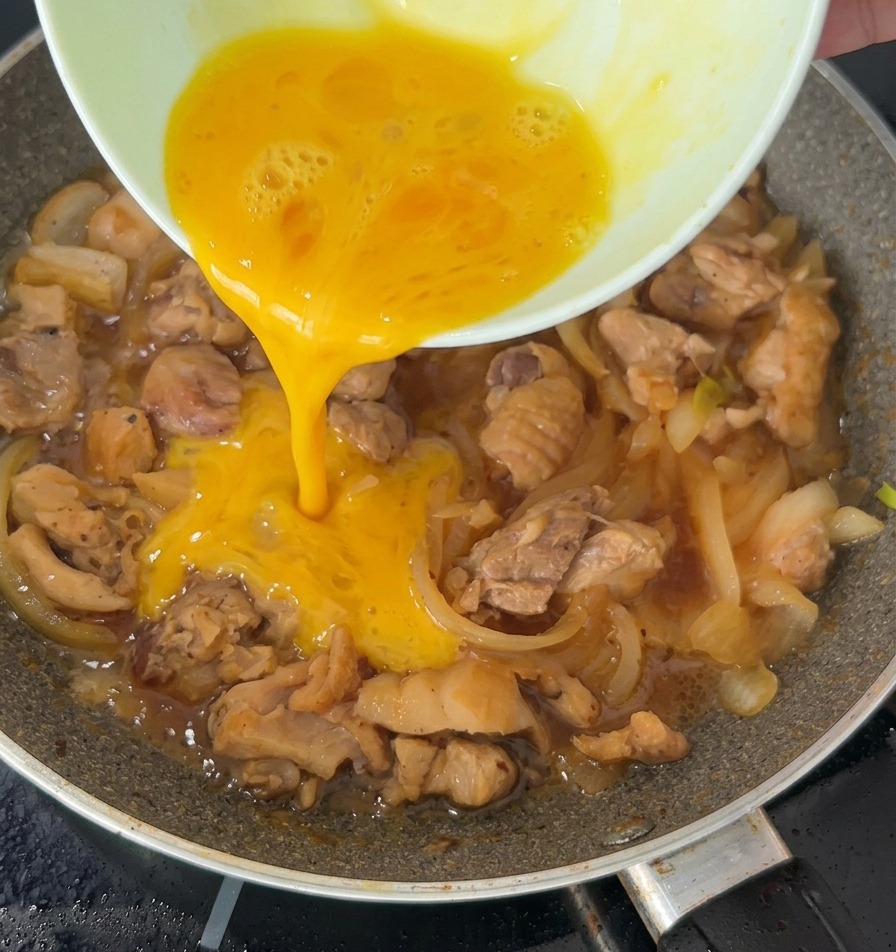 A stream of beaten egg being poured over simmering chicken and onions in a frying pan.
