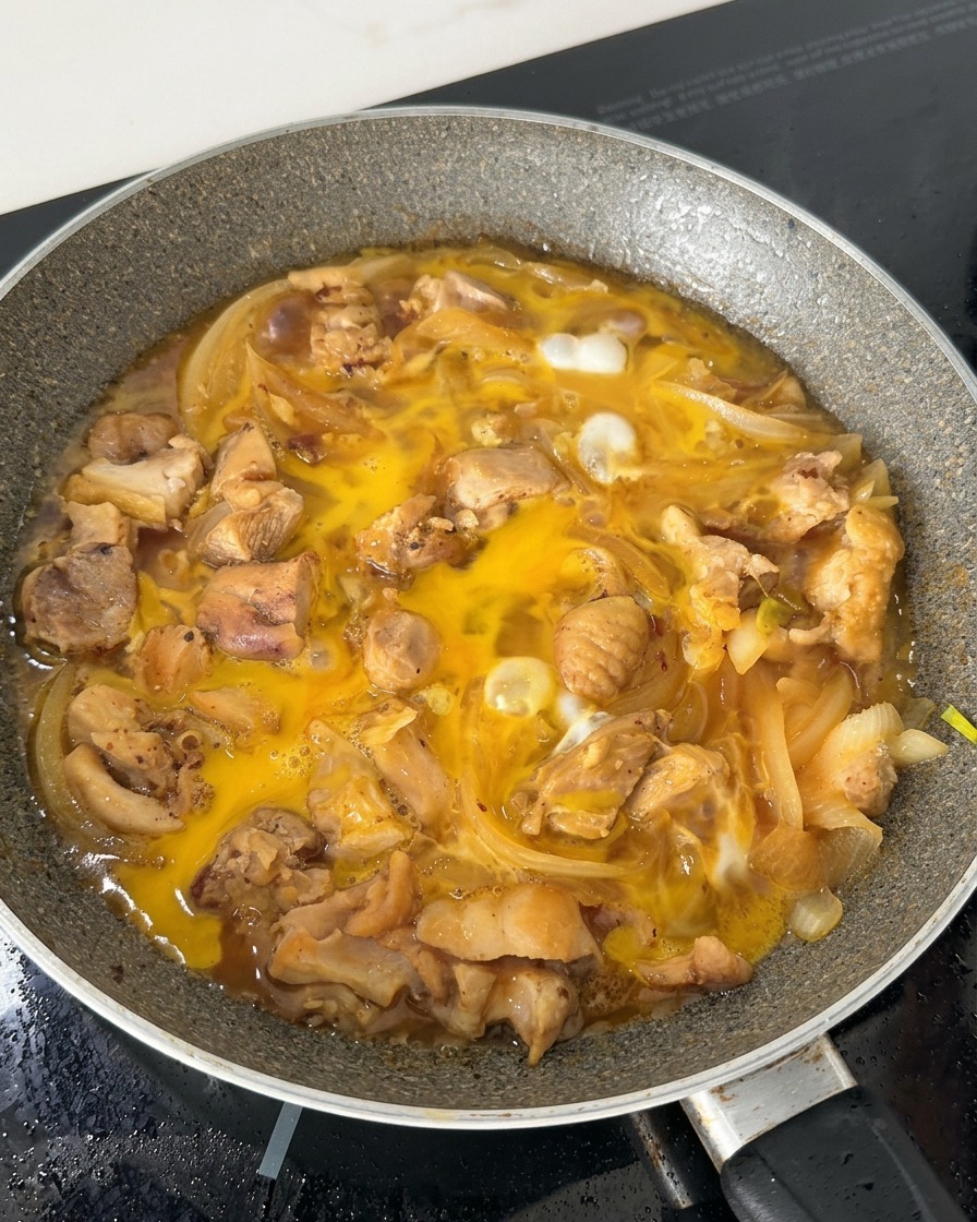 Partially cooked egg mixture bubbling in a skillet with chicken pieces and sliced onions in a soy-based broth.
