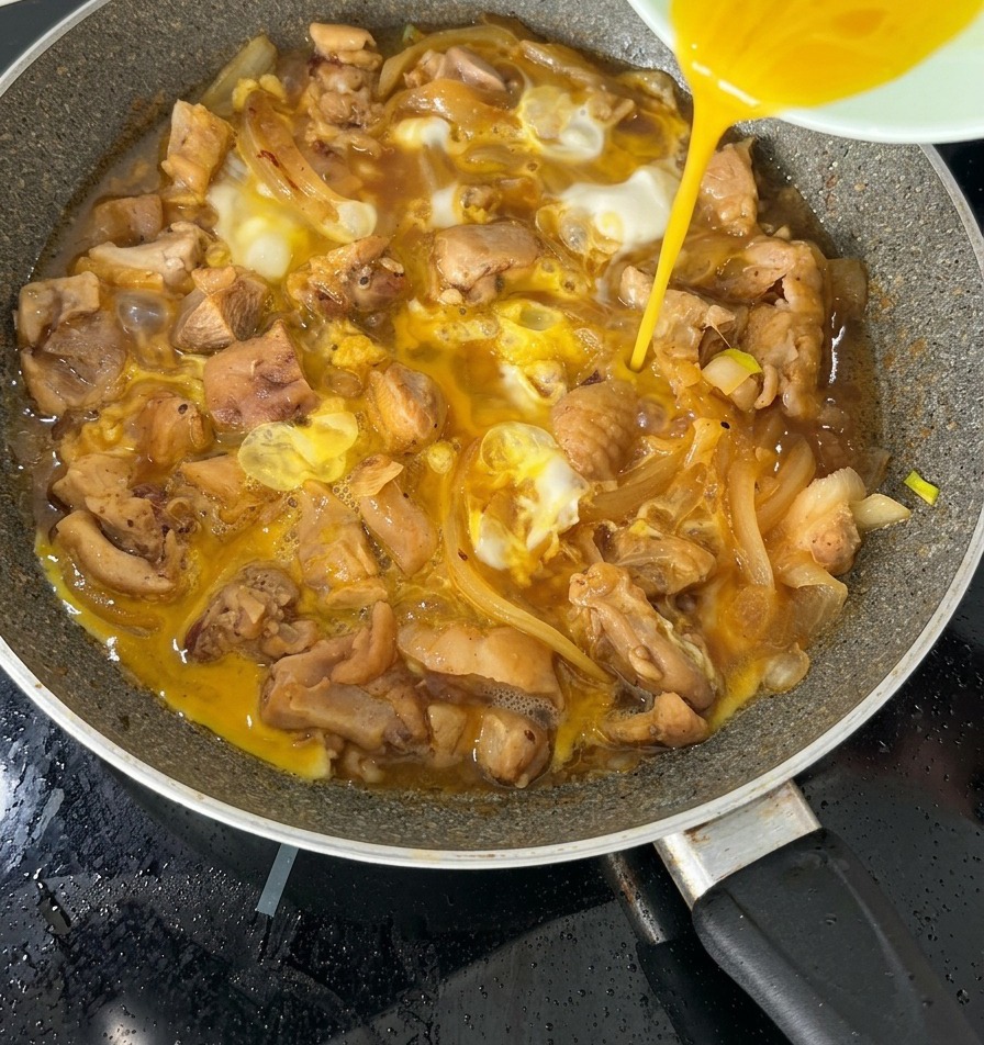 The second half of a beaten egg mixture being poured over a nearly finished Oyakodon in a pan.