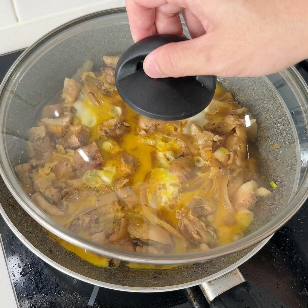A hand placing a glass lid over a skillet of chicken and eggs to steam the final addition of the egg mixture.