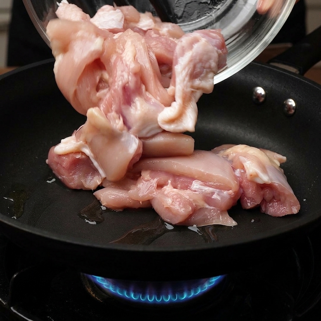 Raw chicken pieces being poured from a glass bowl into a hot frying pan with a blue gas flame visible underneath.