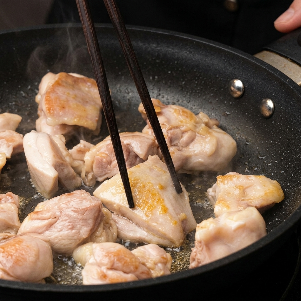 Chicken pieces browning in a pan, being turned with dark wooden chopsticks.