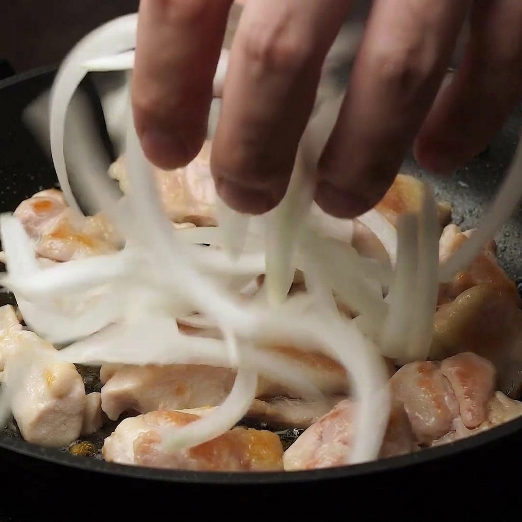 A hand dropping thinly sliced white onions into a pan over partially cooked chicken pieces.