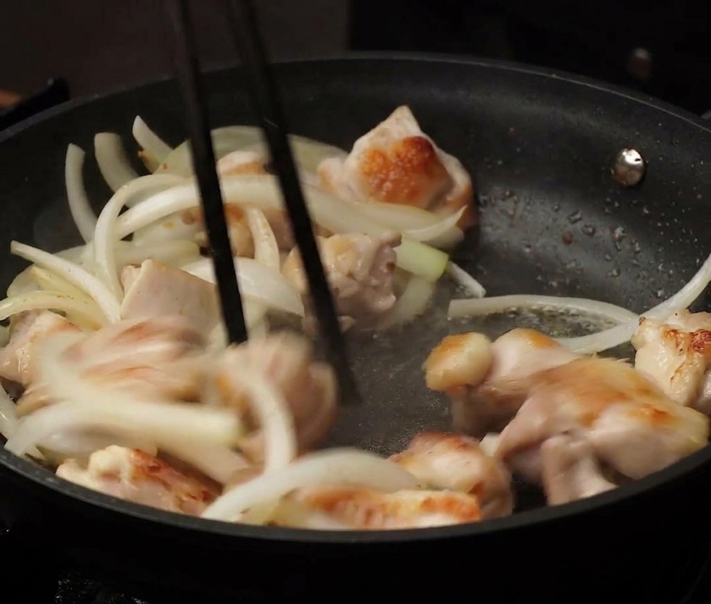 Bite-sized chicken pieces and sliced onions being tossed together in a black non-stick pan using chopsticks.