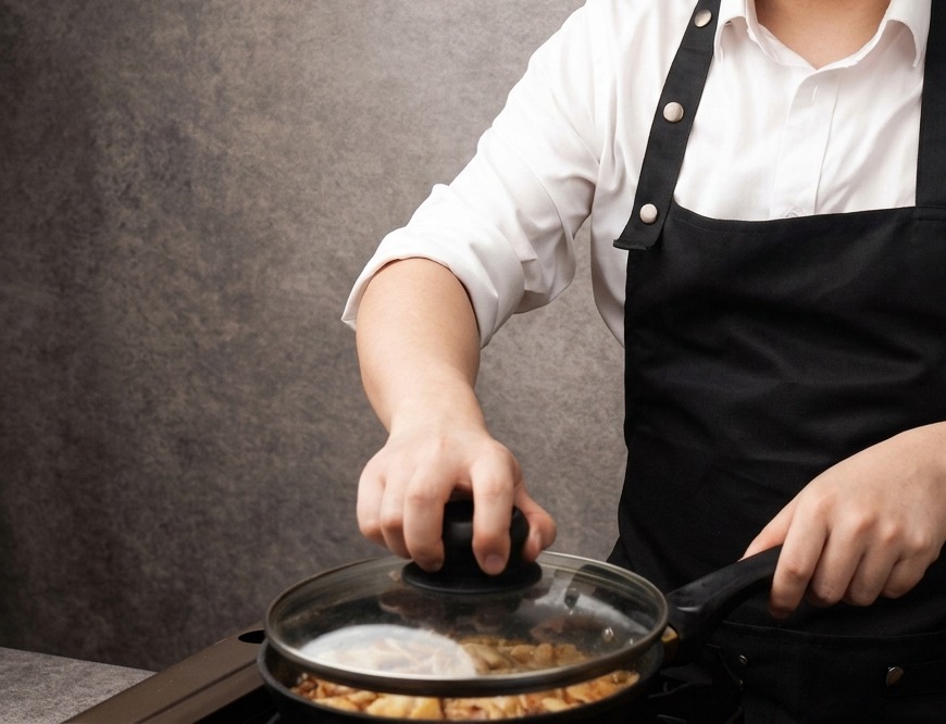 A hand placing a transparent glass lid onto a pan filled with chicken pieces and onion slices simmering in a light brown sauce.