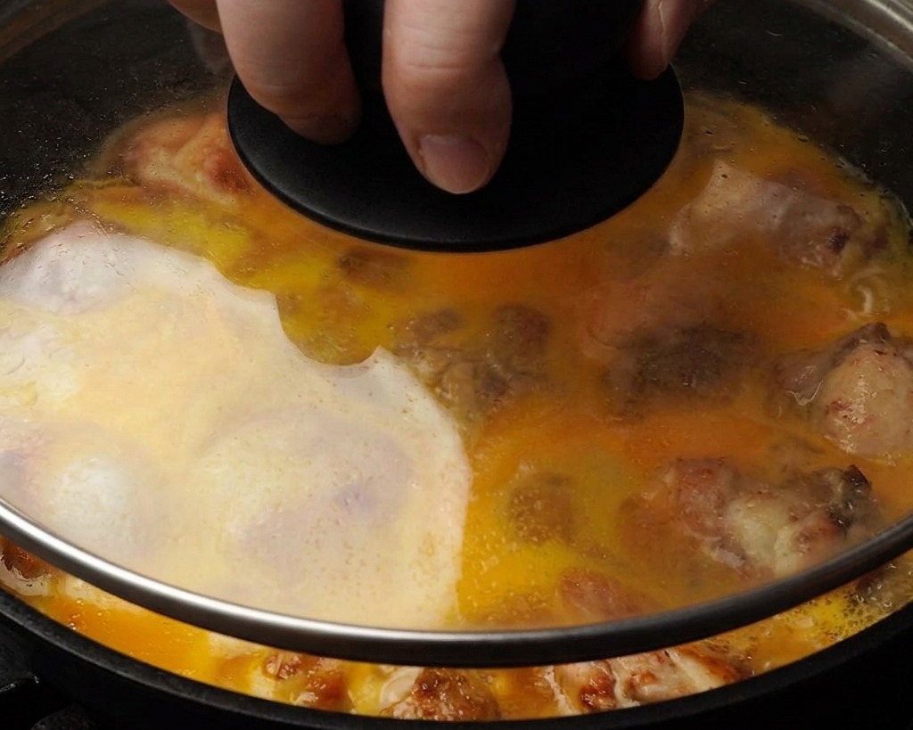 A close-up shot of a pan with a glass lid showing yellow egg liquid partially covering chicken pieces as they steam inside.