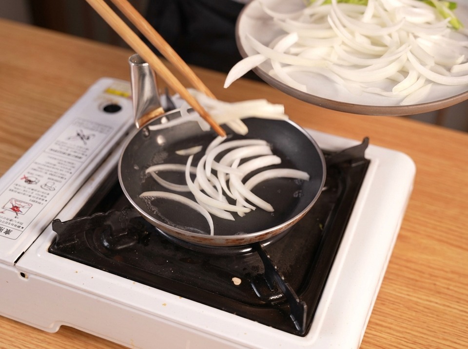 Slices of white onion being added to a small black frying pan on a portable stove from a white plate using wooden chopsticks.