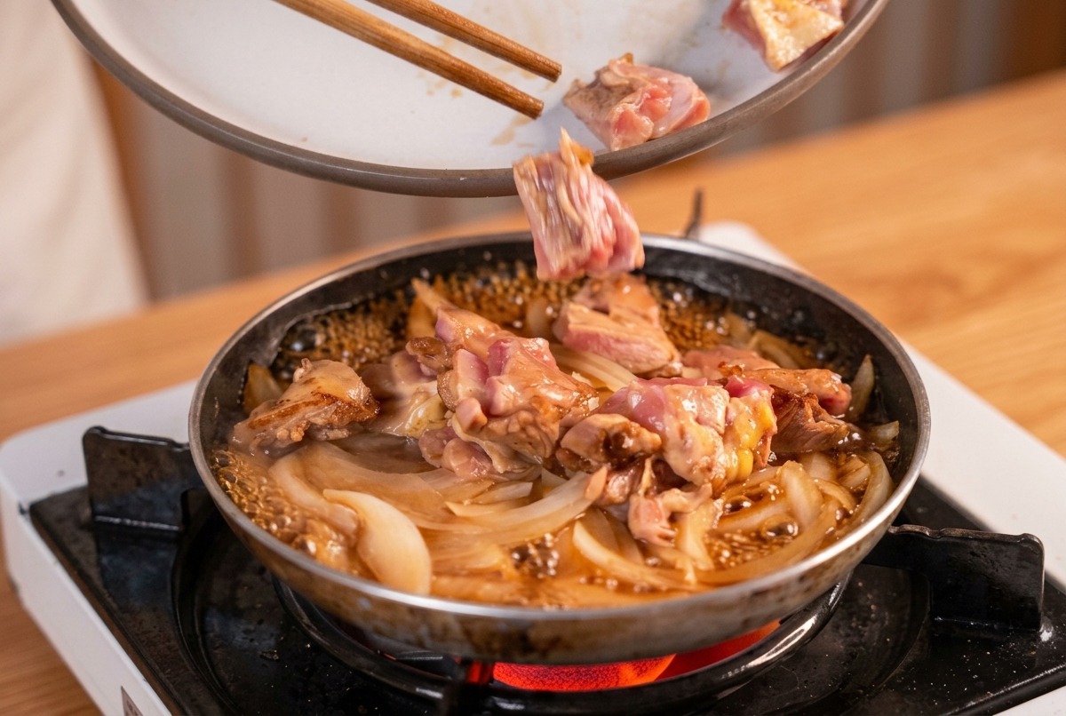 Pieces of seared chicken being added with chopsticks to a pan of simmering onions and dashi broth.