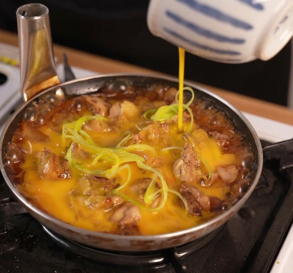 Beaten egg being poured from a blue and white striped bowl over chicken and green onions simmering in a dark sauce.