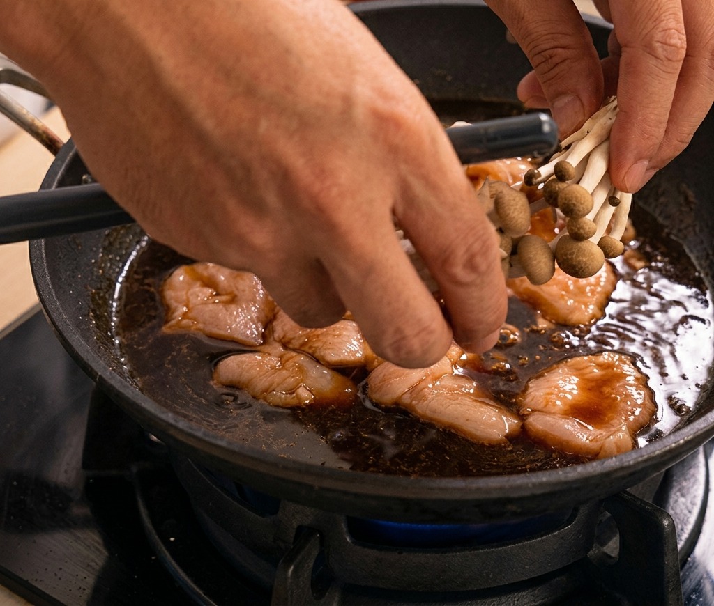 Hands placing a cluster of shimeji mushrooms into a pan of simmering chicken and dark sauce.