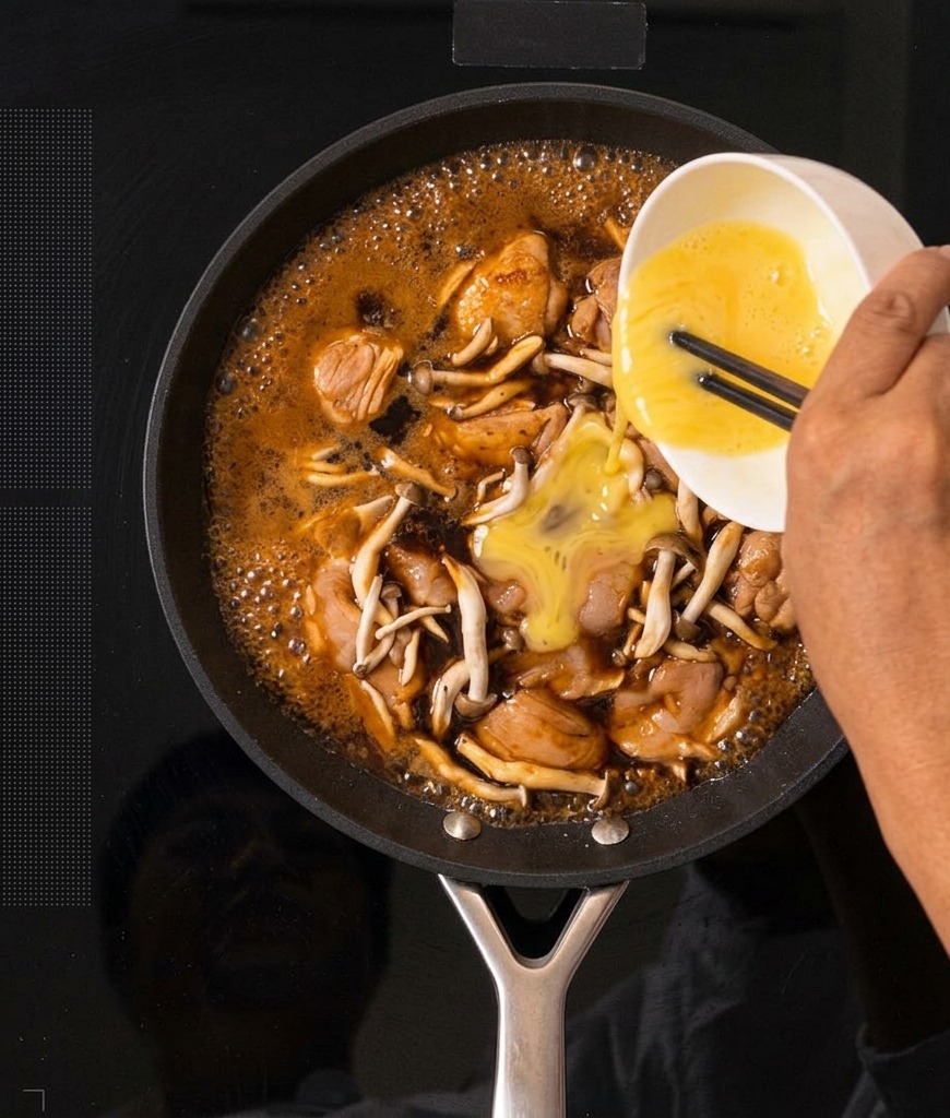 Beaten yellow eggs being poured from a white bowl into a pan of bubbling Oyakodon mixture.