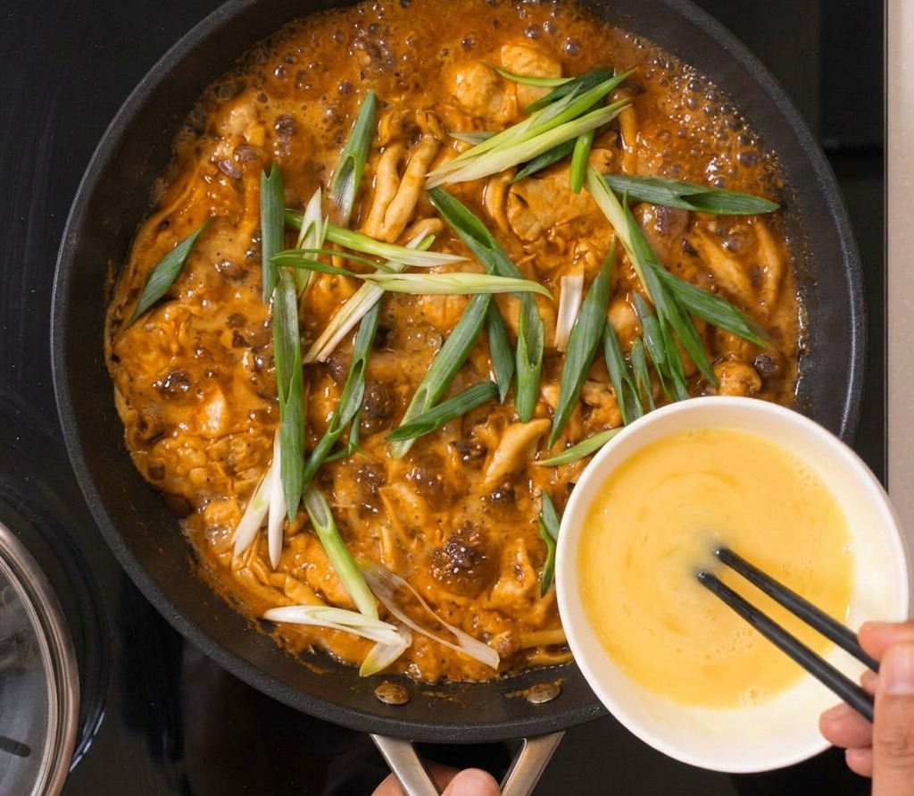 A hand using chopsticks to guide beaten egg from a white bowl into a simmering pan filled with chicken, shimeji mushrooms, and fresh green onions.