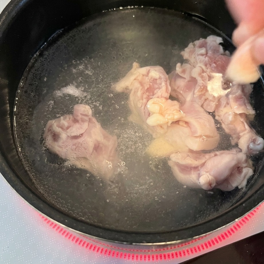 Hands dropping raw chicken chunks into a pot of boiling water on a red induction burner.