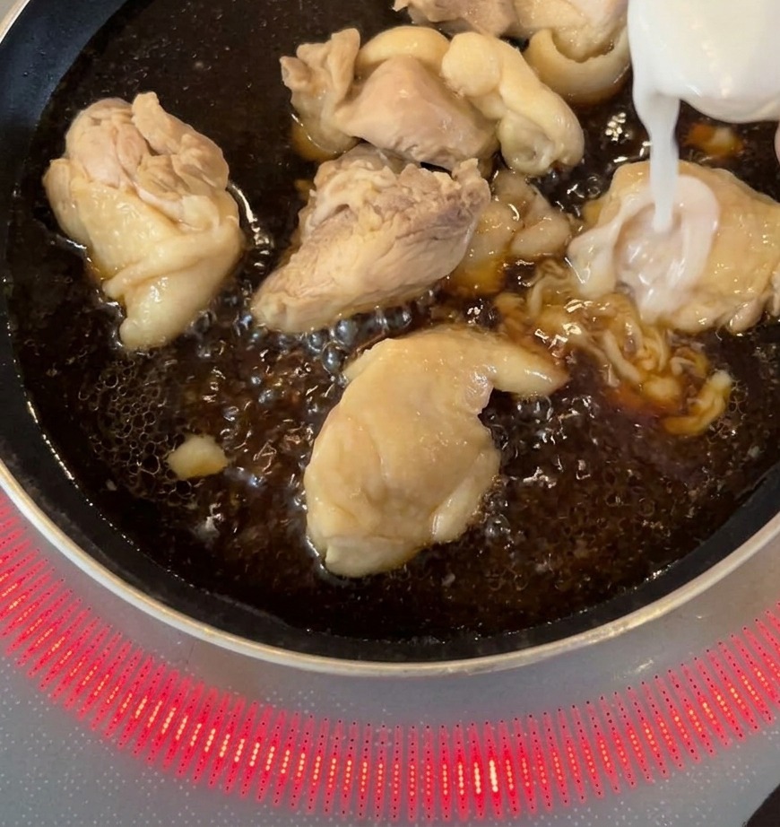 Sliced white onions being added to a pan with chicken pieces simmering in a dark sauce
