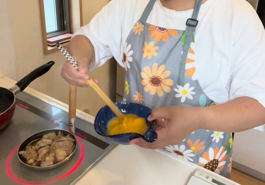 A person using wooden chopsticks to whisk two eggs in a deep blue ceramic bowl, with a pan of simmering chicken visible on the stove nearby.