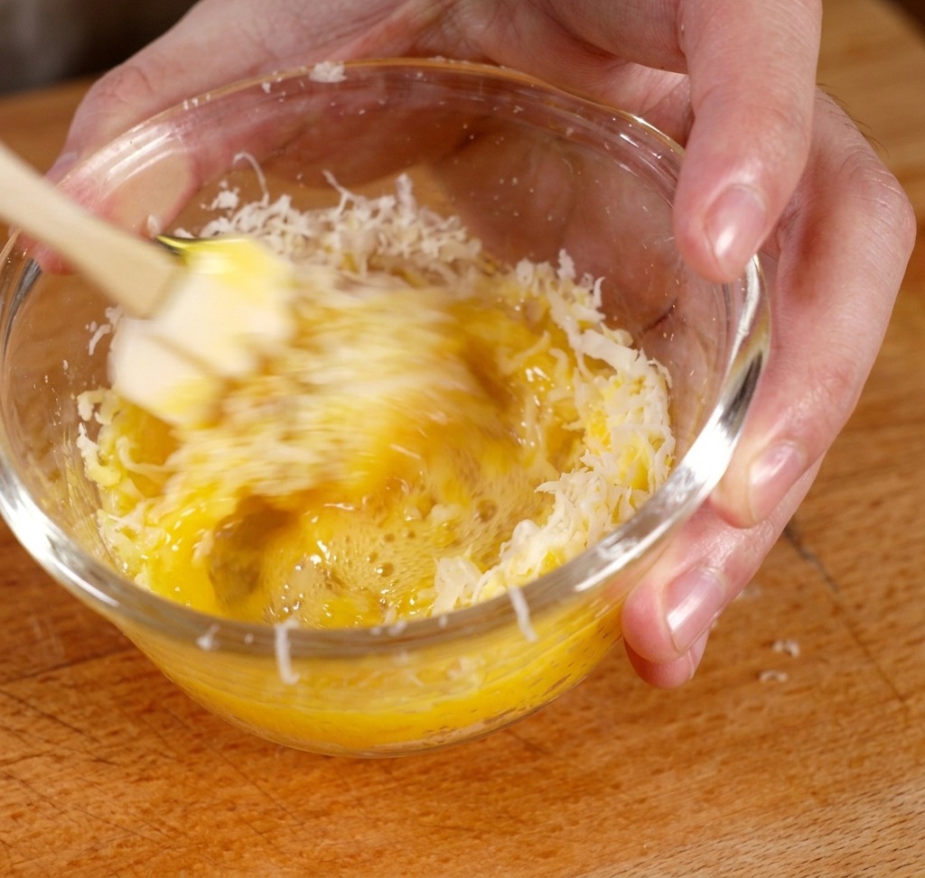 Whisking egg yolk and grated cheese together in a glass bowl to form a thick yellow paste.