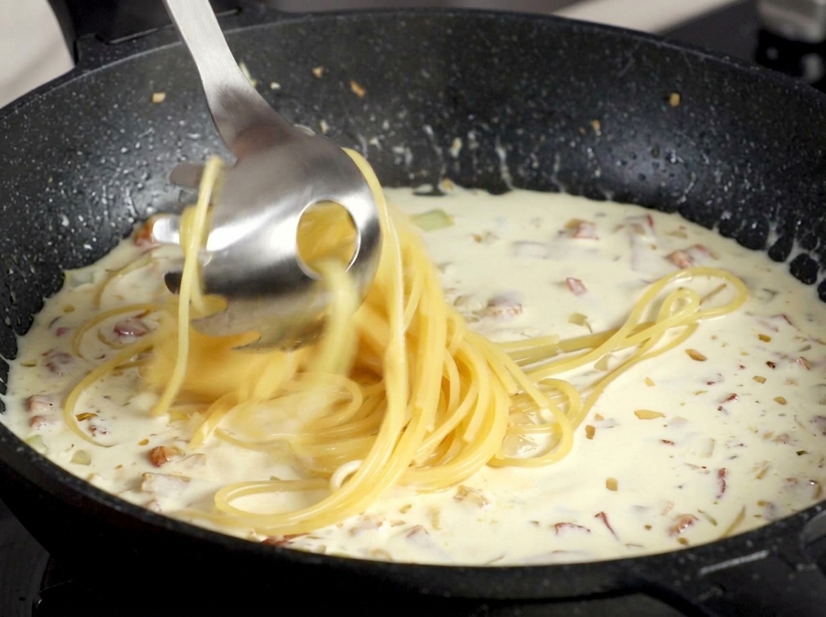 Spaghetti being lifted with a metal pasta spoon into a creamy white bacon sauce.
