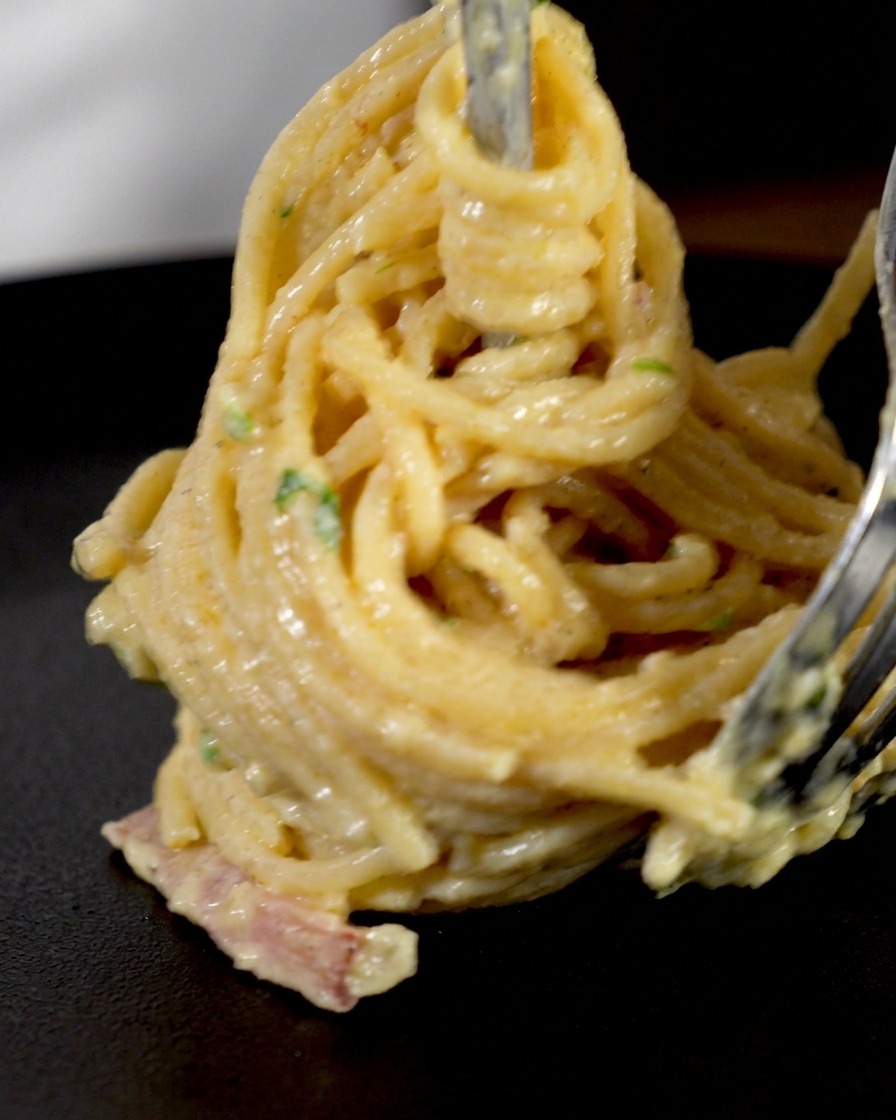 Creamy spaghetti and bacon pieces being tightly twirled around a long metal carving fork to form a neat vertical nest for plating.