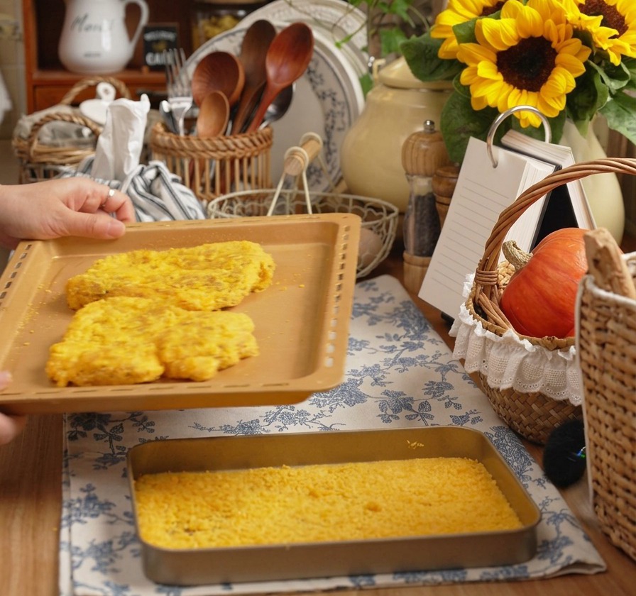 Hands holding a baking sheet with two large breaded pork cutlets in a warm kitchen setting.