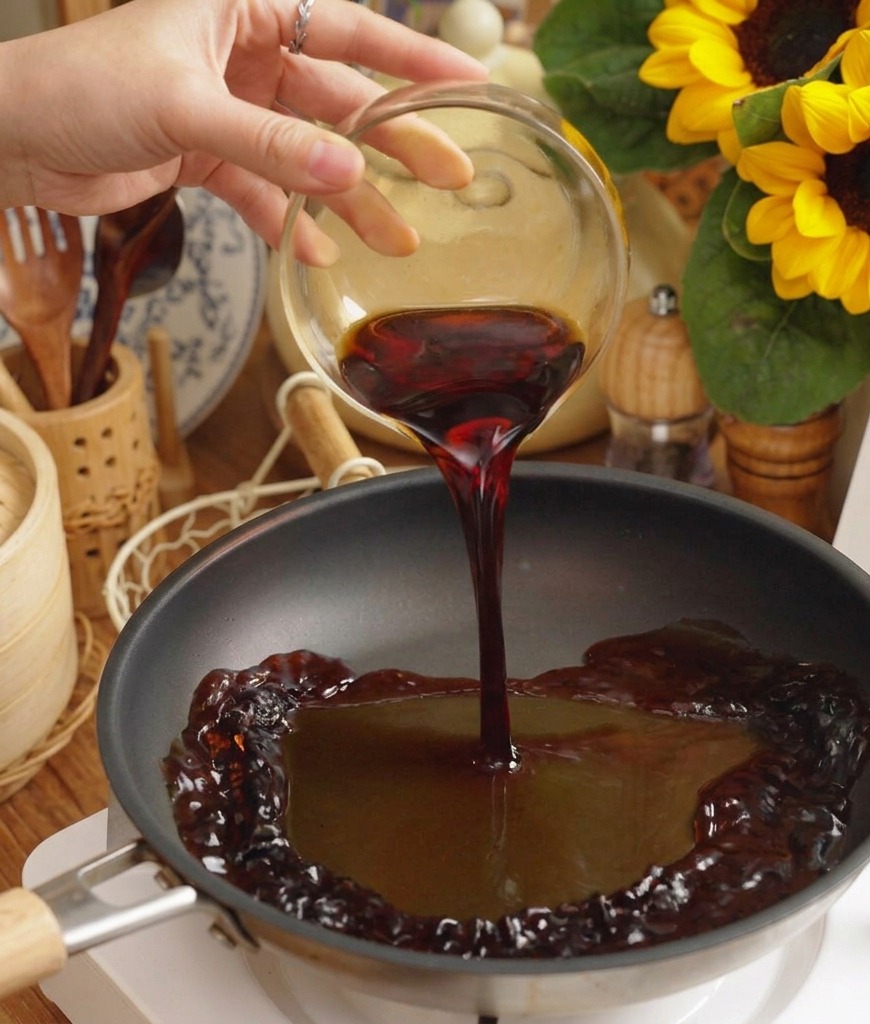 Dark savory sauce being poured from a glass bowl into a black frying pan on a white electric cooktop.