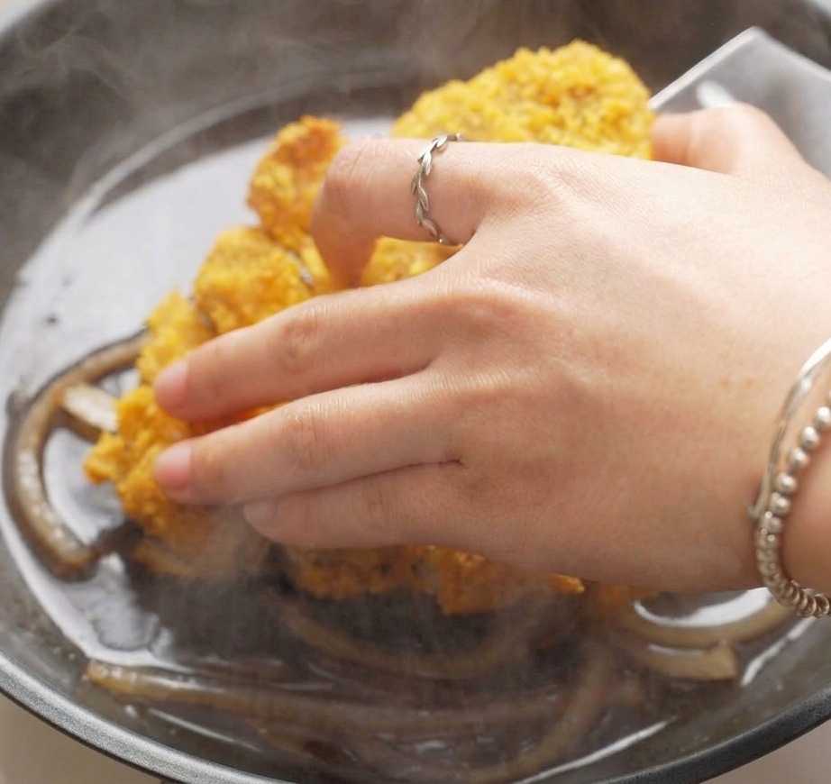 A hand placing slices of golden-brown breaded pork cutlet into a frying pan filled with simmering sliced onions and a dark savory sauce.