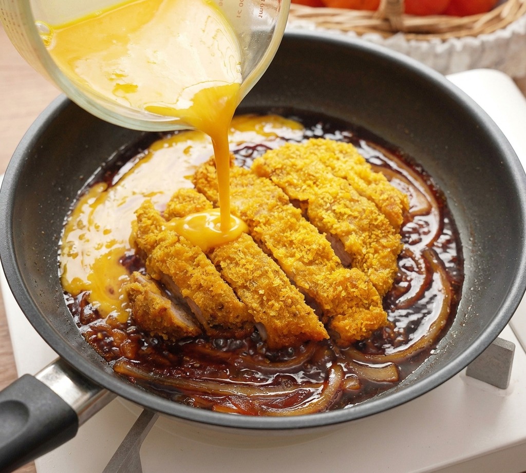 Yellow beaten eggs being poured from a glass measuring cup into a pan containing a sliced fried pork cutlet and onions.