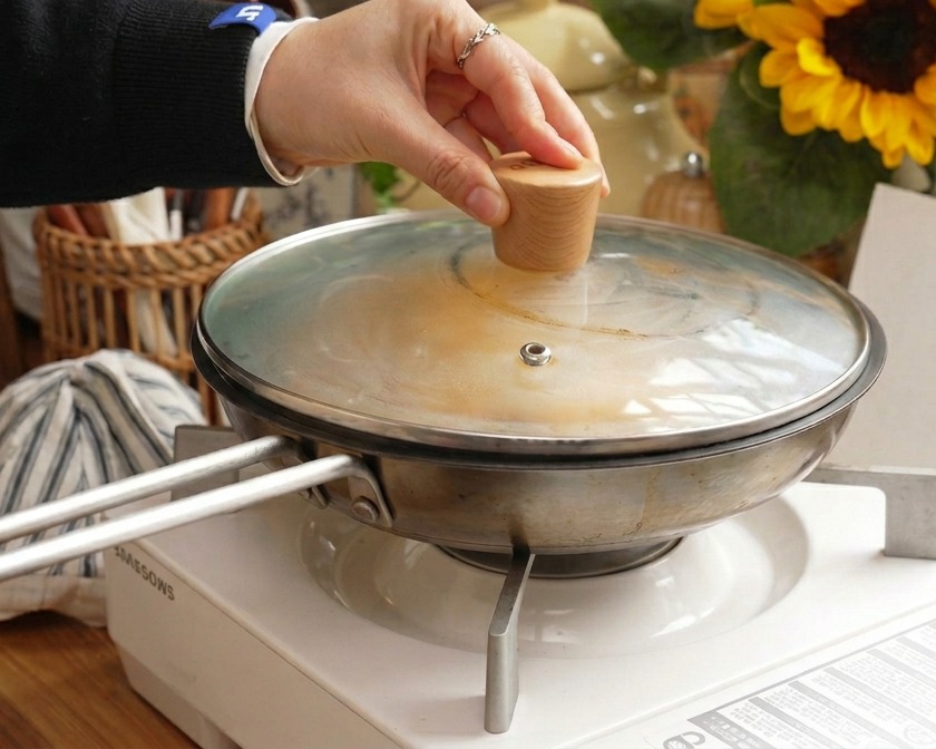 A hand holding the wooden knob of a glass lid as it is placed onto a stainless steel pan to steam the pork and egg dish.