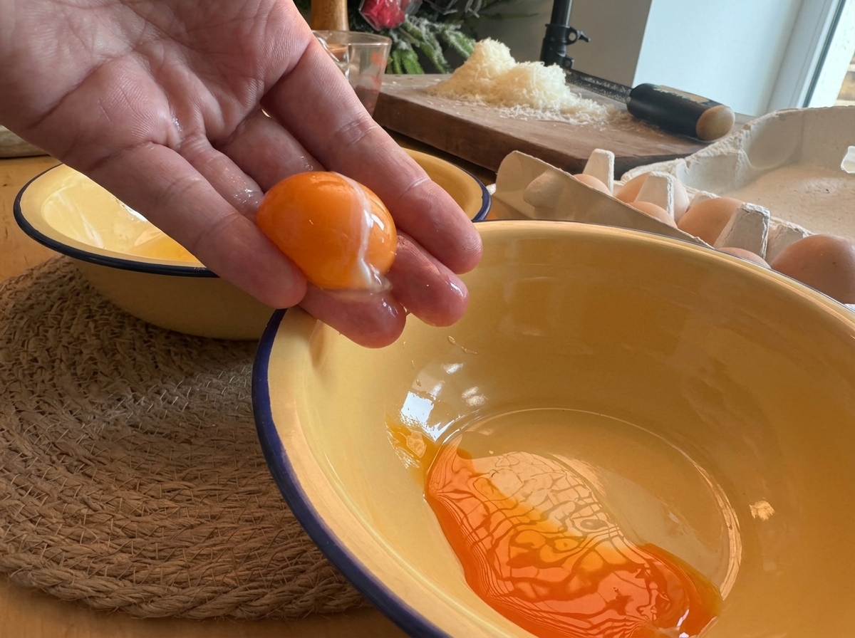 A hand separating a bright orange egg yolk over a yellow enamel bowl.
