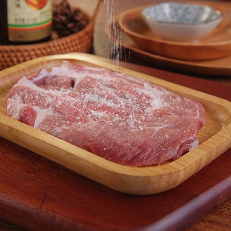 Salt being sprinkled from a hand onto a raw pork cutlet on a wooden serving tray.