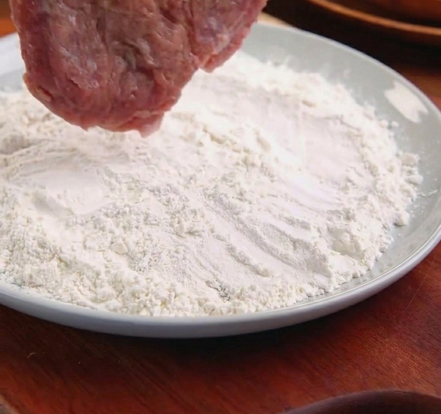 A pork cutlet held by metal tongs being lowered into a plate of white flour.