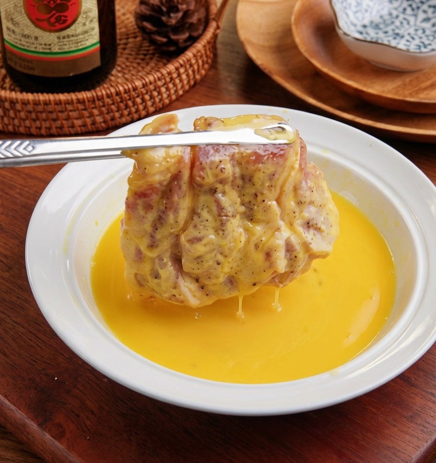 A floured pork cutlet being dipped into a white bowl filled with yellow egg wash.