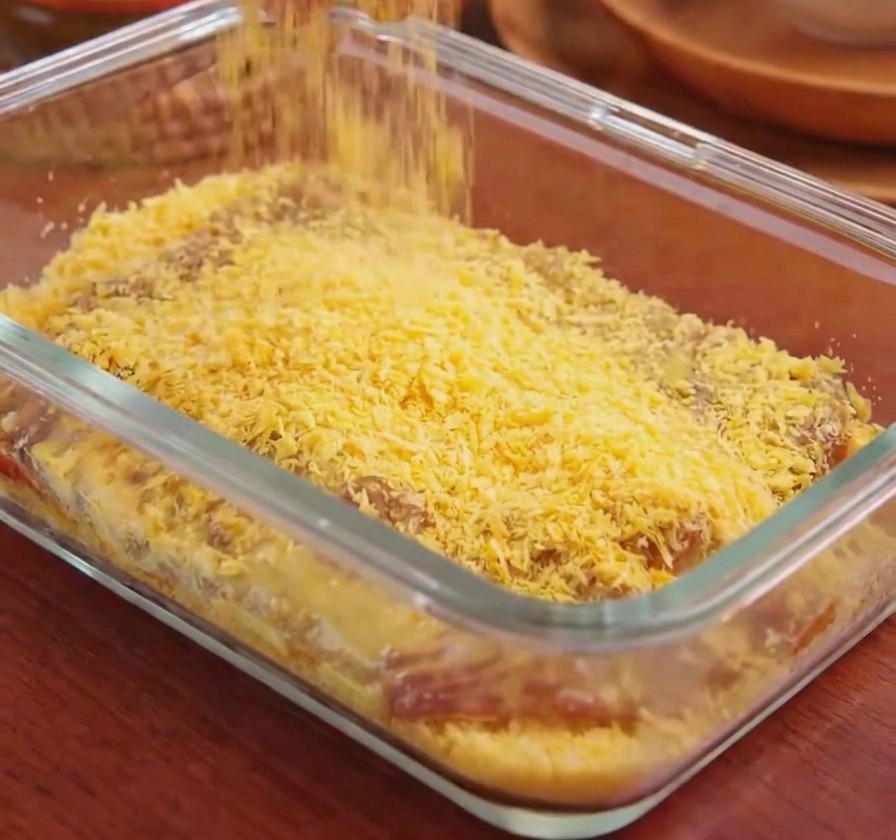 Hand sprinkling golden-yellow breadcrumbs over a pork cutlet in a glass container.