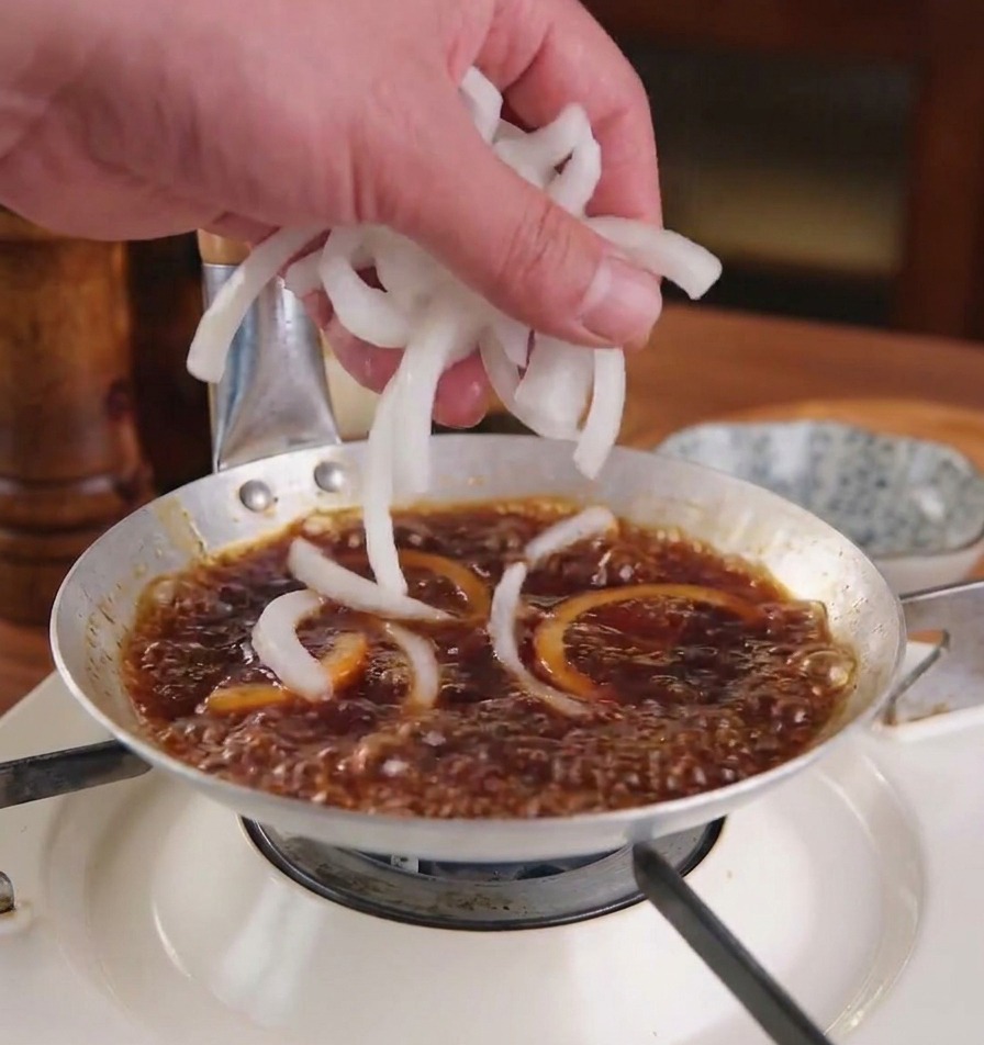 A hand adding fresh white onion slices into a bubbling dark brown sauce in a shallow metal cooking pan.