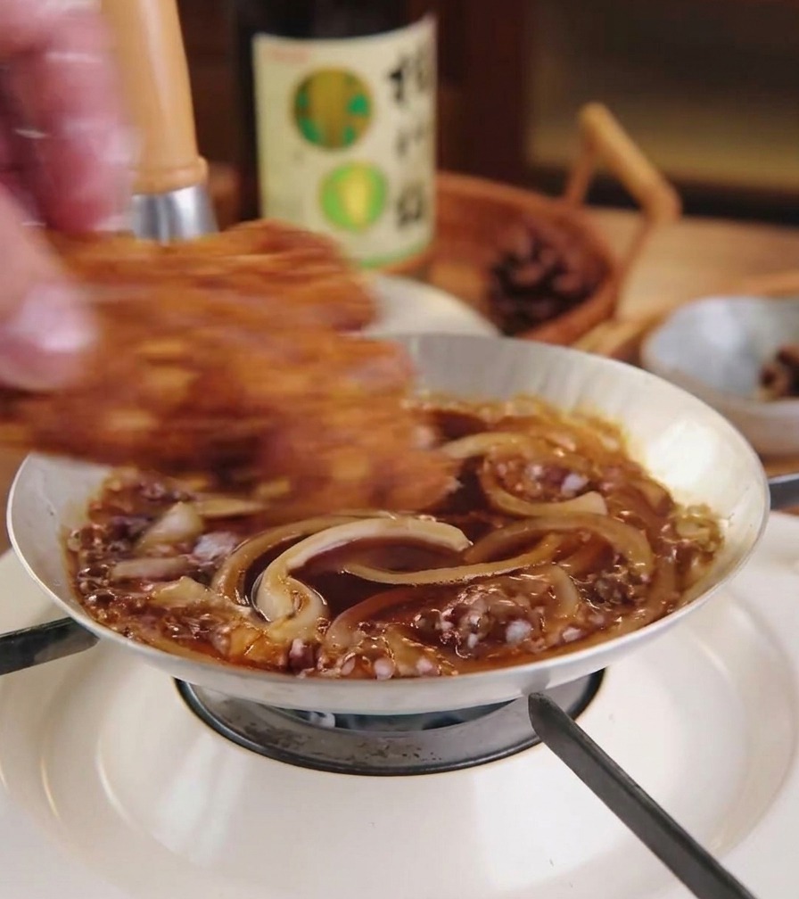 A fried, golden-brown pork cutlet being placed on top of a layer of simmering onions and sauce in a small pan.