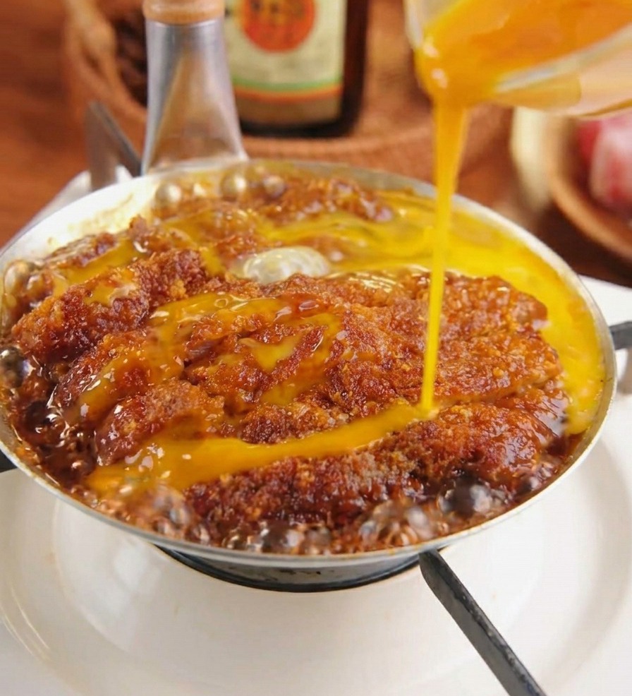 Beaten yellow egg liquid being poured over a breaded pork cutlet simmering in a dark sauce with onions.