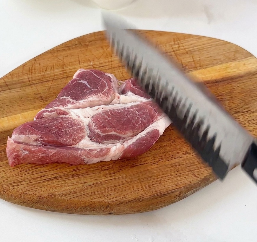 A hand using the back of a chefs knife to tenderize a raw pork cutlet on a wooden board.