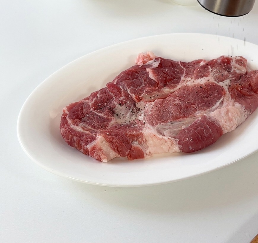 Black pepper being ground over a raw pork cutlet resting on a white ceramic plate.