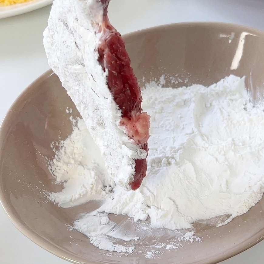 A pork cutlet being coated in white flour in a tan ceramic bowl.