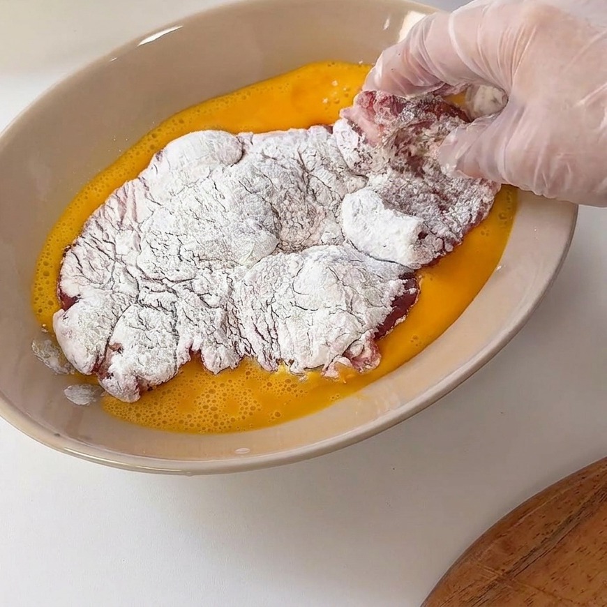 A floured pork cutlet being dipped into a shallow bowl filled with yellow whisked egg wash.