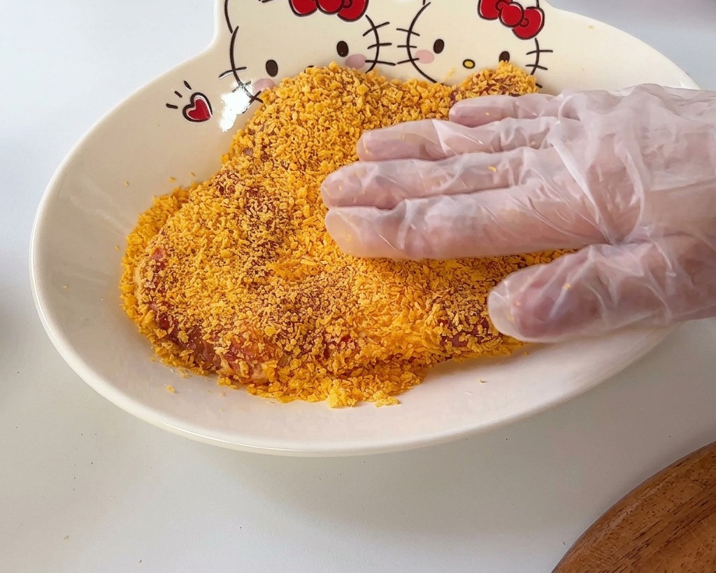 A hand pressing a pork cutlet into a layer of orange breadcrumbs in a bowl with a Hello Kitty design.