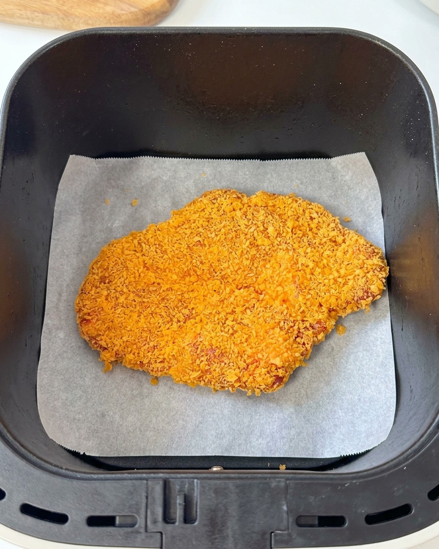 A breaded pork cutlet resting on parchment paper inside a black air fryer basket.