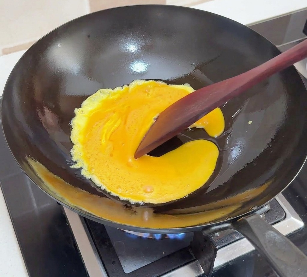 Beaten eggs cooking in a black wok, being pushed toward the center with a dark spatula to create soft folds.