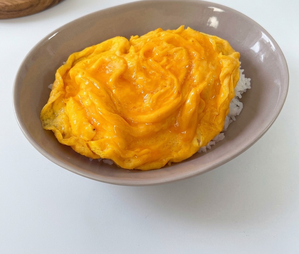 A fluffy, soft-cooked yellow omelet resting on top of a bed of white rice in a shallow grey bowl.