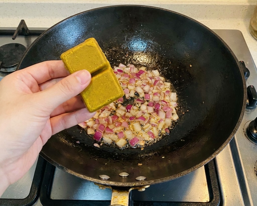A person holding two brown curry blocks over a wok containing sautéed red onion pieces.