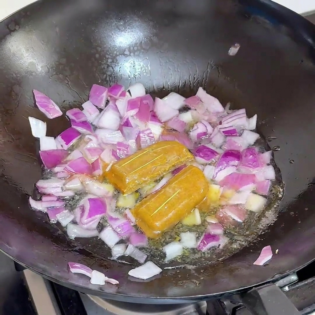 Water being added to a black wok filled with red onions and two whole curry blocks.