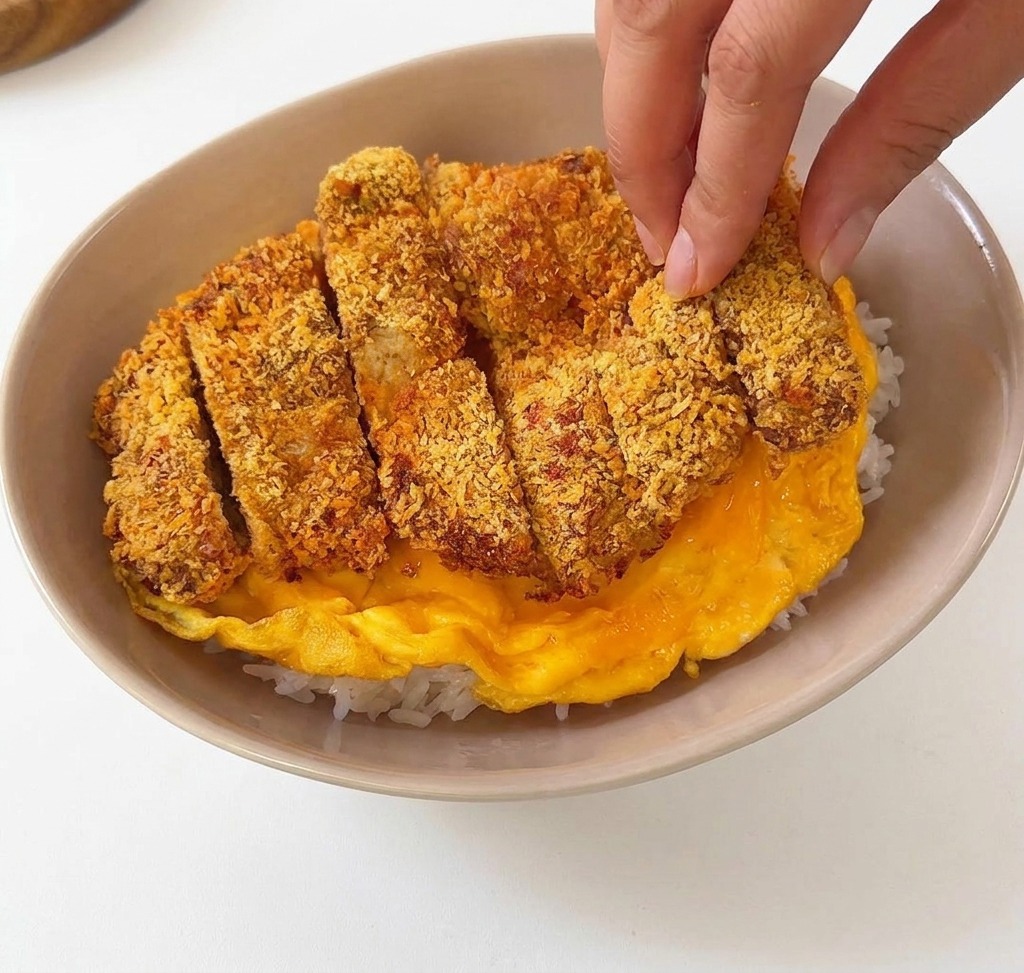 Sliced breaded pork cutlet being placed on top of a soft yellow omelet inside a beige ceramic bowl of white rice.